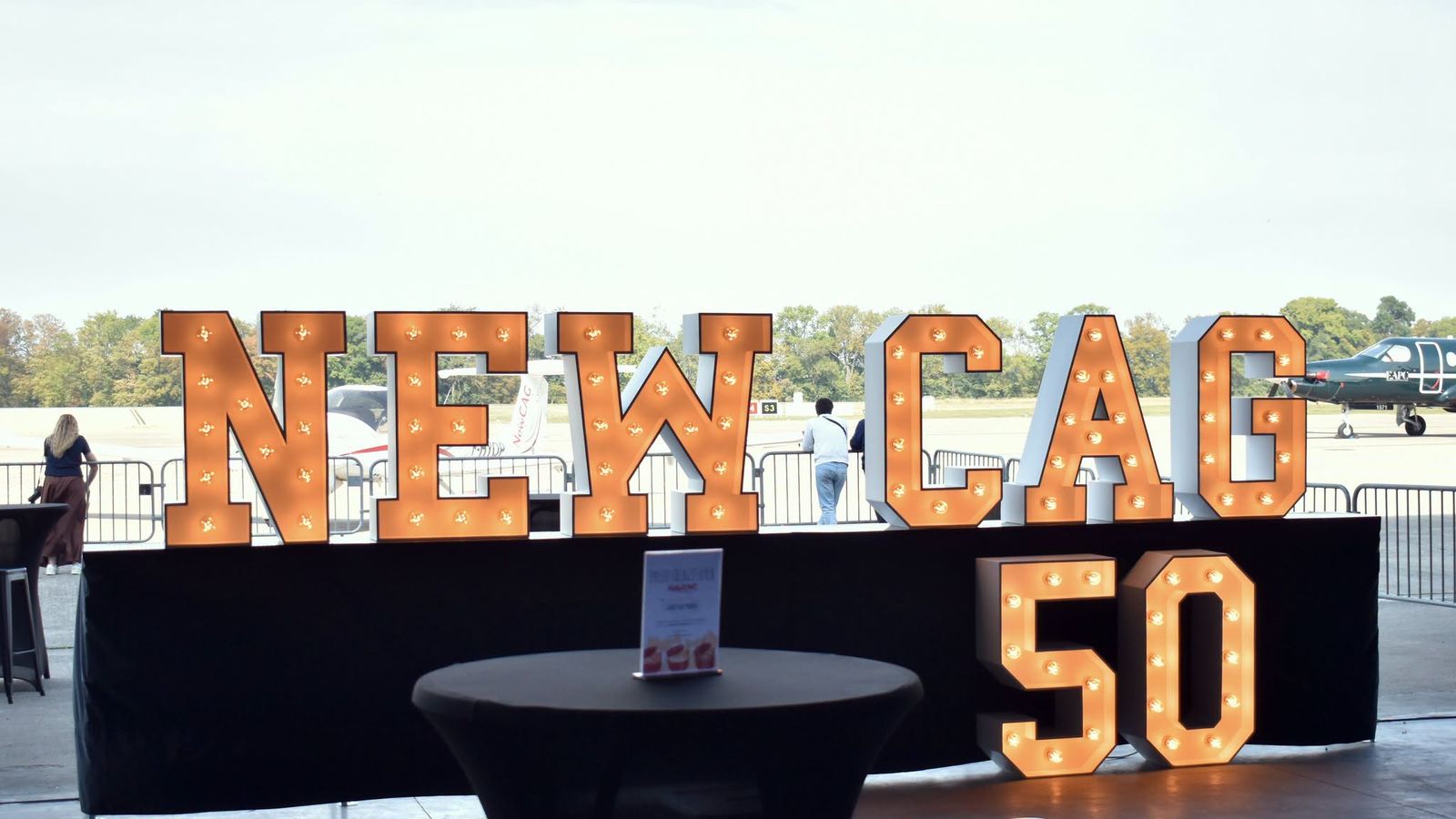 Les lettres lumineuses « NEWCAG 50 » dans le hangar, devant le tarmac et les avions de l’école